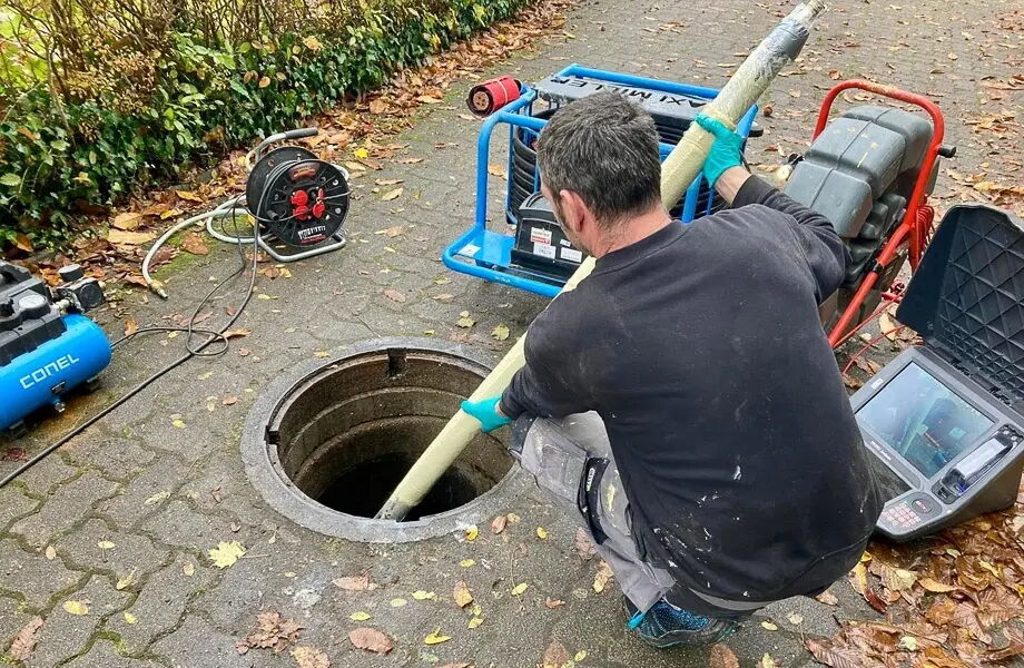 Ein Arbeiter in blauer Handschuhen steckt einen Schlauch in einen Gully auf einem gepflasterten Weg. Neben ihm stehen Maschinen und Kabel. Der Boden ist mit Laub bedeckt, und im Hintergrund ist eine Hecke zu sehen.