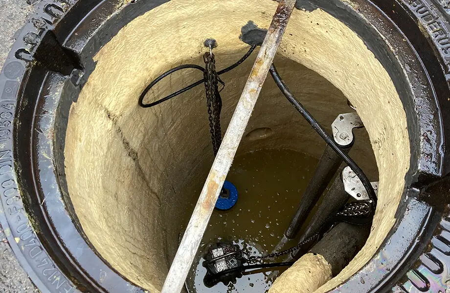 Ansicht eines offenen Kanalschachts mit rostbraunem Metallrand. Im Inneren befindet sich trübes Wasser und verschiedene Metallketten hängen herab. Ein blauer Schwimmer ist im Wasser sichtbar, ebenso wie mehrere Metallstangen und Rohre.