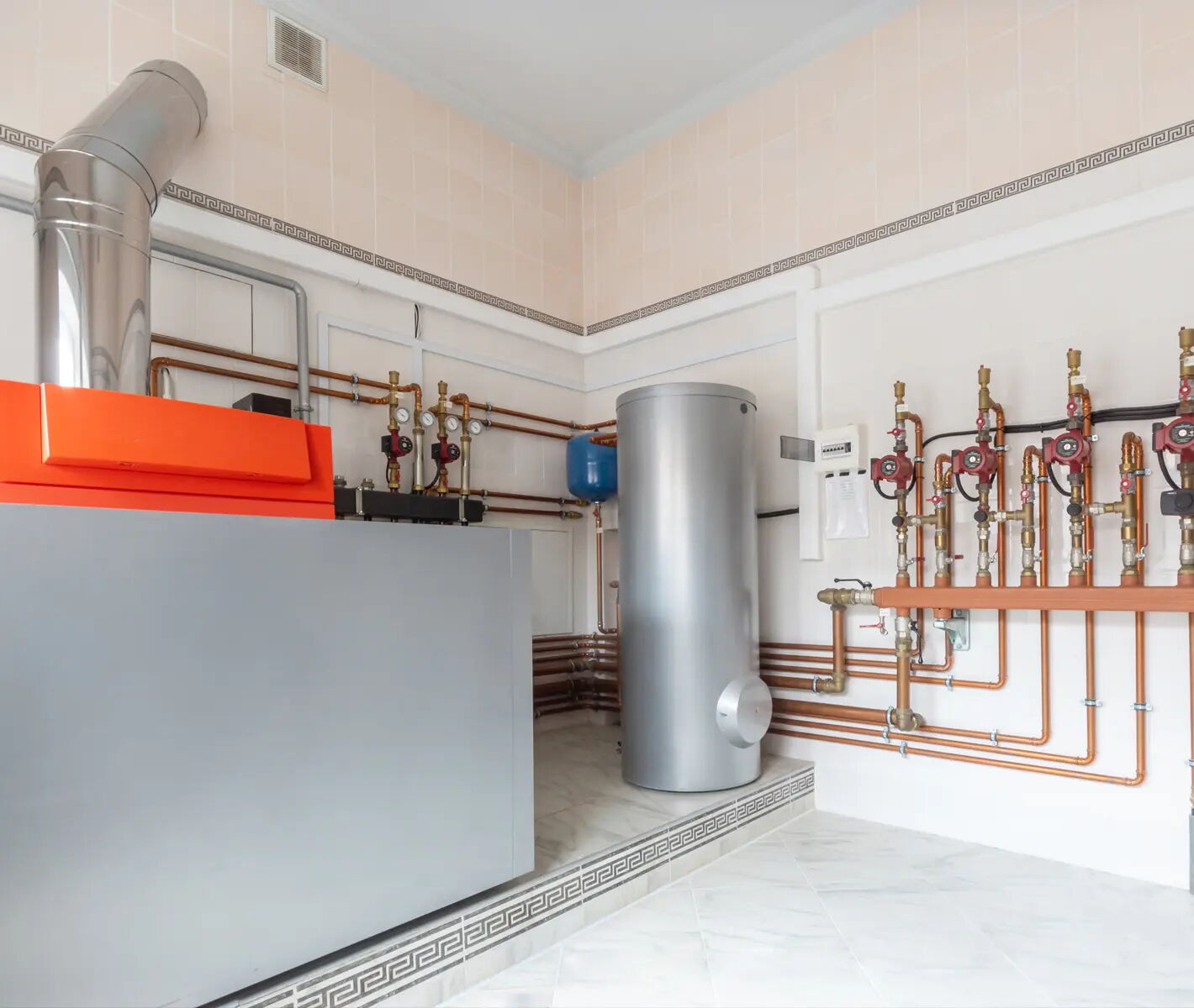 Ein Technikraum mit Heizungsanlage, bestehend aus einem großen silbernen Tank, orangefarbenem Heizsystem, Rohren und Ventilen an der Wand. Der Raum hat helle Wände und einen gefliesten Boden.