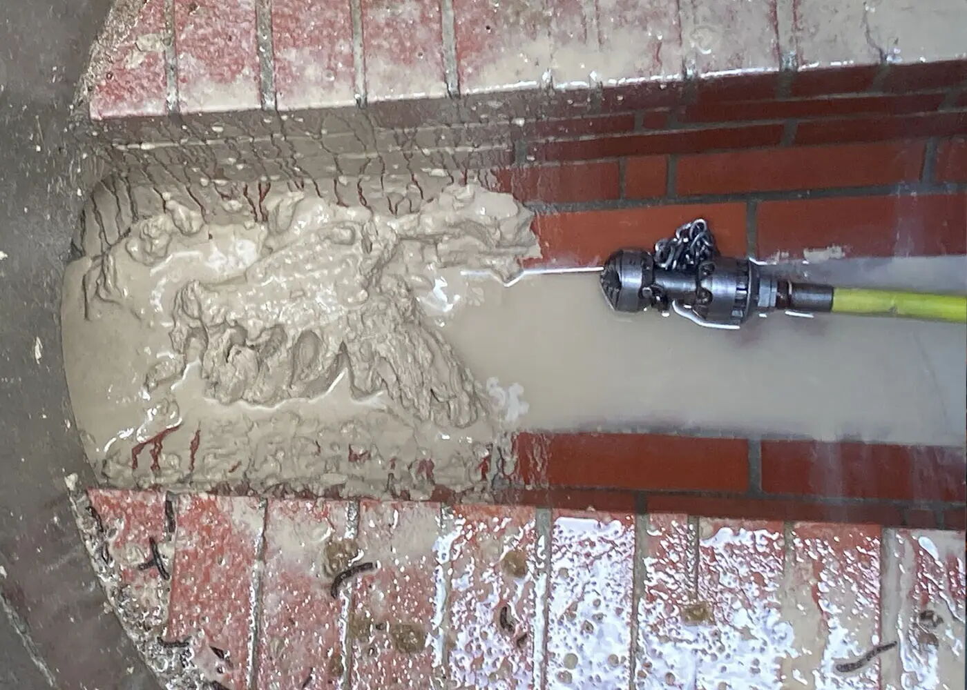 Ein Wasserstrahl aus einem Schlauch reinigt schmutzige, rotbraune Ziegelsteine. Der Bereich links des Strahls ist mit braunem Schlamm bedeckt, während rechts die Steine sauber und nass sind.