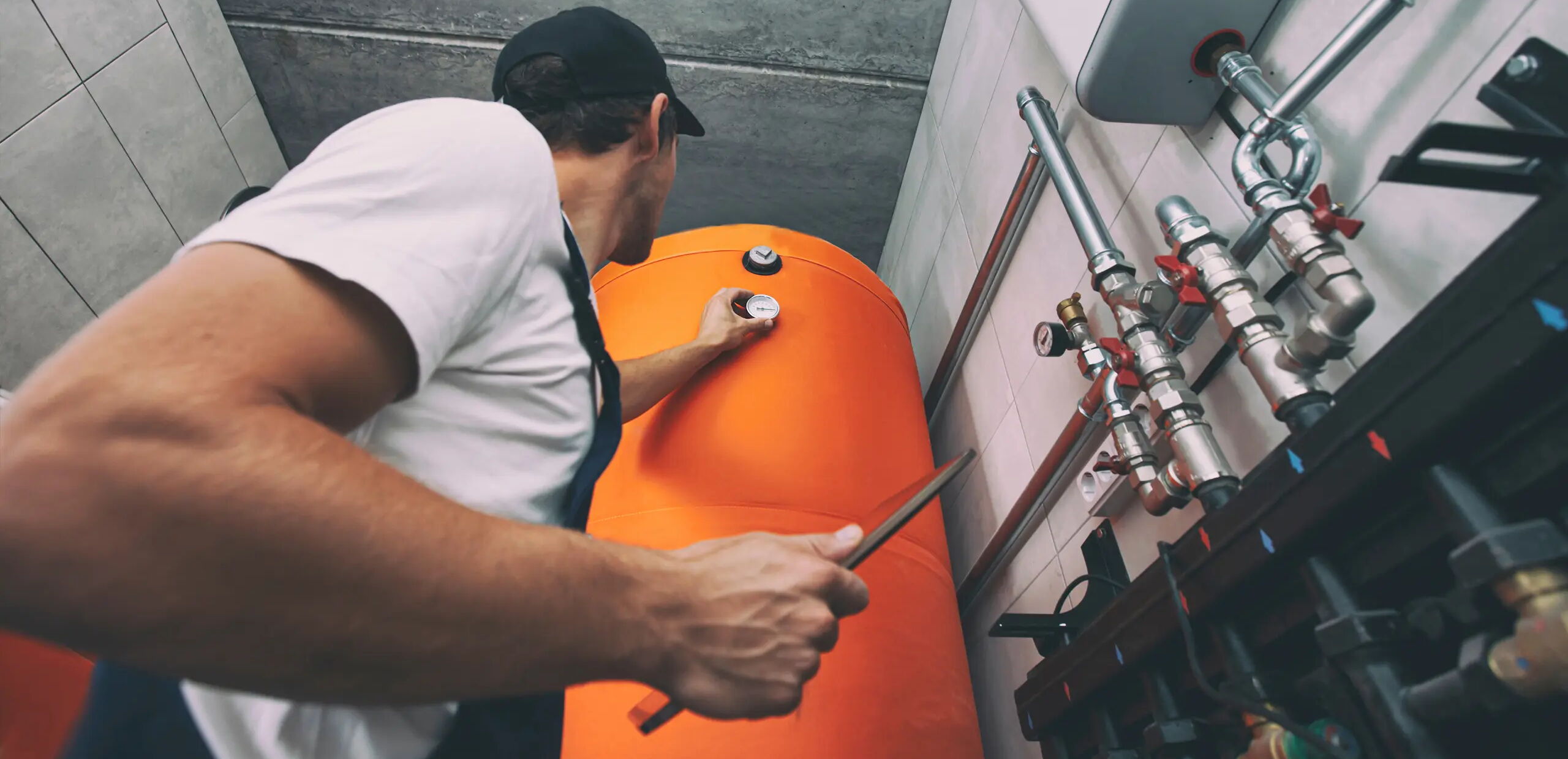 Ein Mann in Arbeitskleidung prüft eine orangefarbene Druckanzeige an einem großen Tank in einem Technikraum. Er hält ein Werkzeug in der Hand. Rohre und Ventile sind im Hintergrund sichtbar.