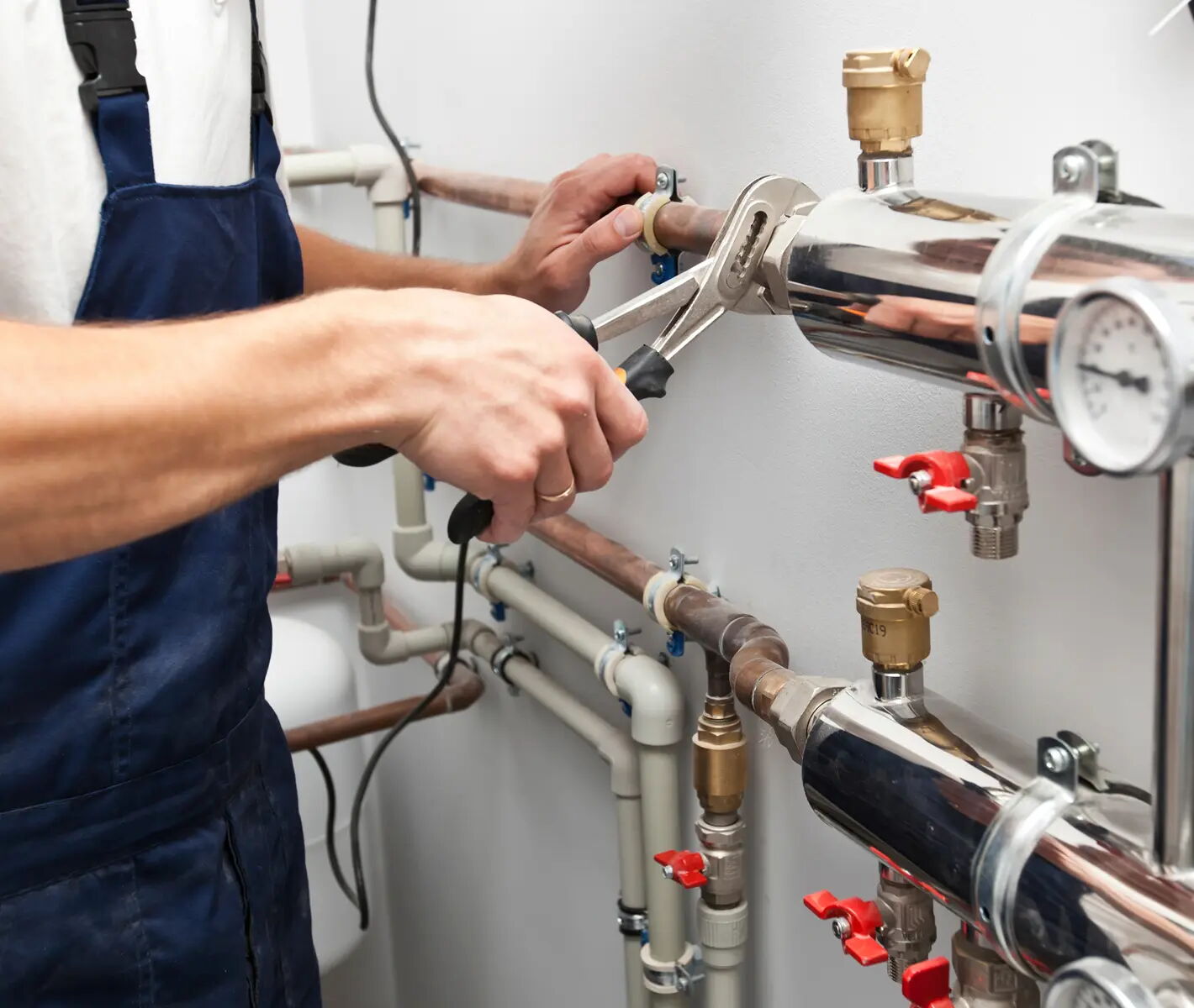 Person in Arbeitskleidung benutzt einen verstellbaren Schraubenschlüssel, um an einem Rohrsystem mit Ventilen und einem Druckmanometer zu arbeiten. Verschiedene Rohrleitungen sind an der Wand montiert.