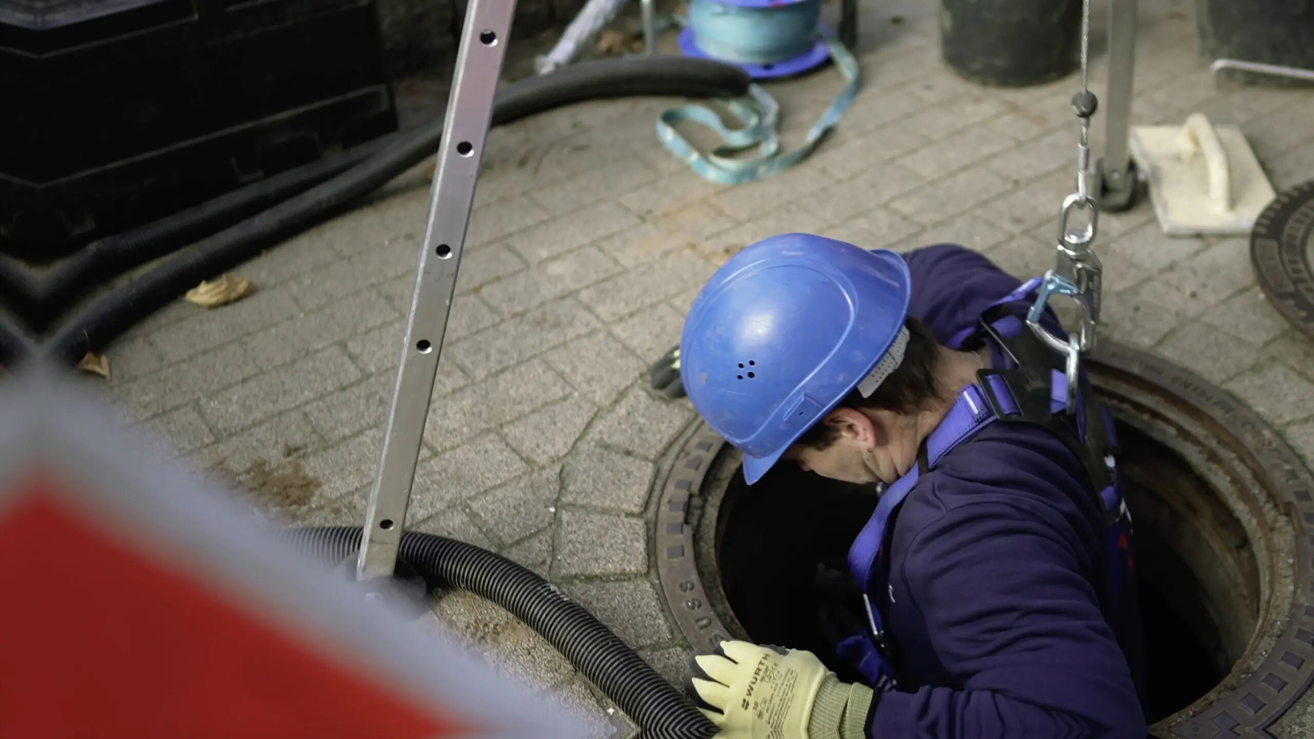 Eine Person in Sicherheitskleidung mit blauem Helm und Handschuhen steigt in einen Gullyschacht. Eine Leiter steht daneben, und einige Ausrüstungsgegenstände liegen auf dem gepflasterten Boden in der Nähe.