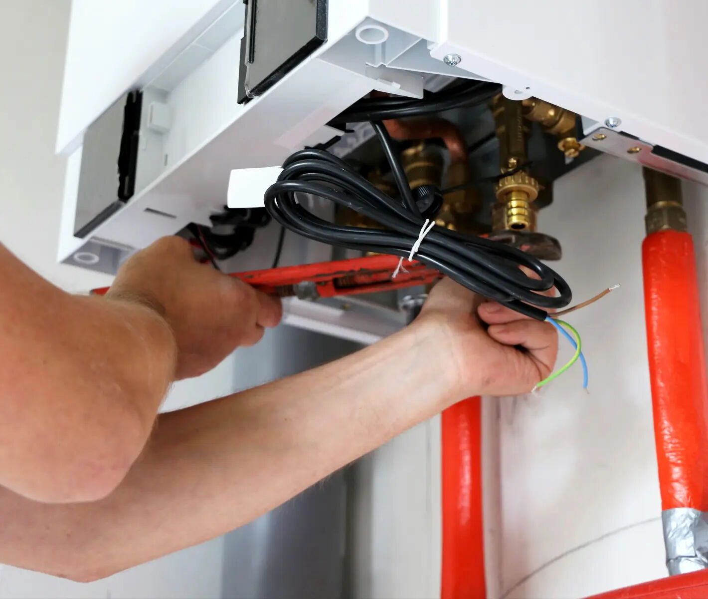 Ein Paar Hände arbeitet an der Unterseite eines Boilers. Die linke Hand hält ein Werkzeug, die rechte Hand hält Kabel. Der Boiler ist an der Wand montiert, darunter sind rote Rohre sichtbar.