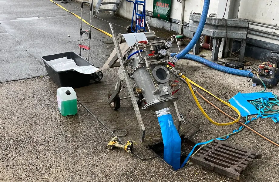 Eine Maschine mit Metallgehäuse pumpt eine blaue Röhre in einen Gully. Daneben stehen ein schwarzer Behälter, ein grüner Kanister und mehrere Schläuche. Der Boden ist nass, und im Hintergrund sind diverse Geräte zu sehen.