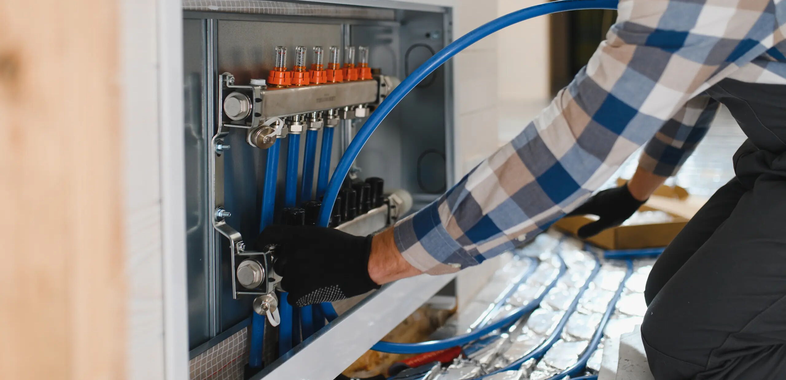 Eine Person mit kariertem Hemd und schwarzen Handschuhen arbeitet an einer Heizungsinstallation. Blaue Röhren sind an eine Verteilungseinheit angeschlossen. Der Boden ist mit reflektierenden Materialien bedeckt.
