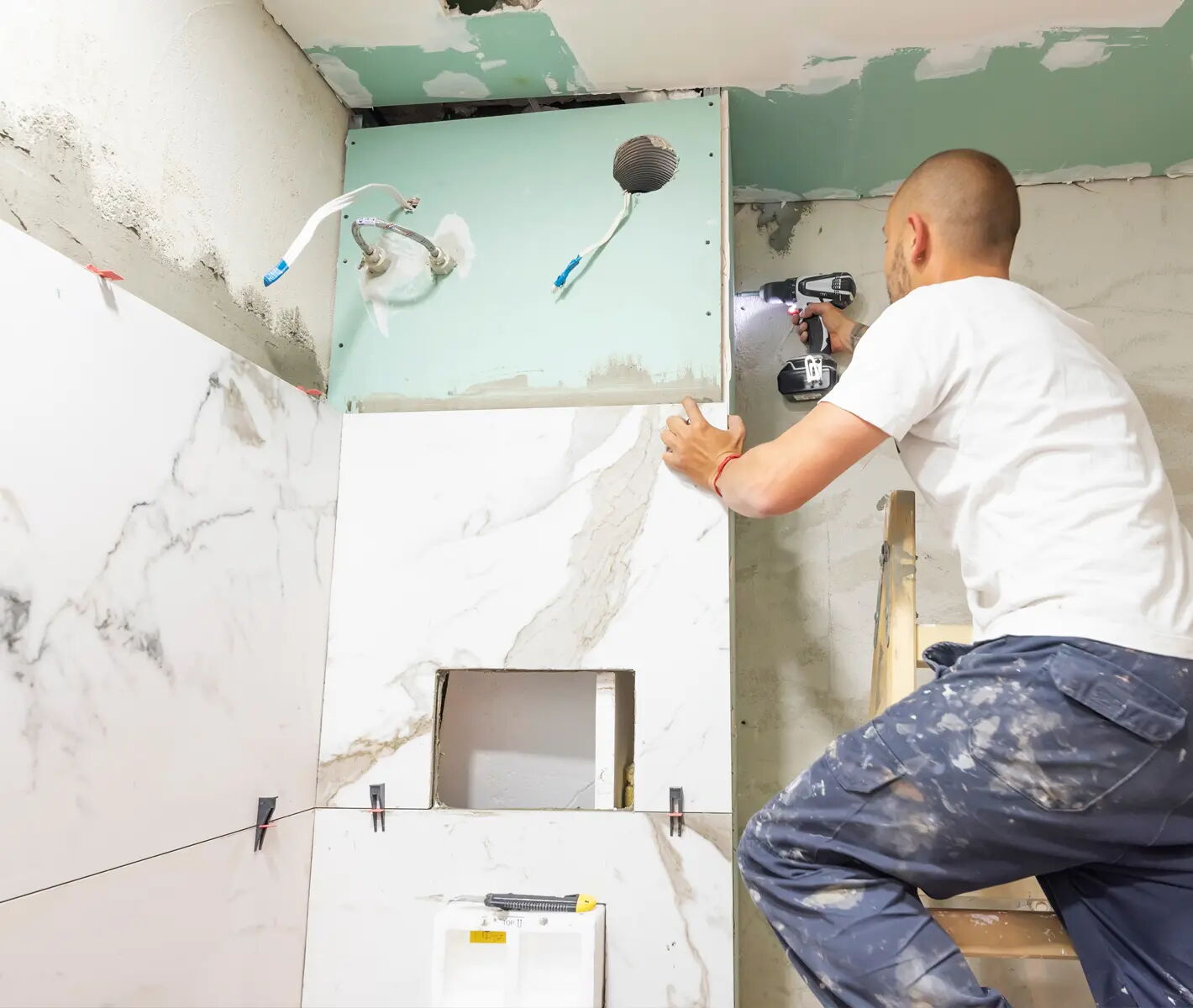 Ein Handwerker auf einer Leiter befestigt mit einem Akkuschrauber eine Trockenbauplatte an einer Badezimmerwand. Die Wand hat Marmorfliesen, und es sind Leitungen für Dusche und Wasserhahn sichtbar.