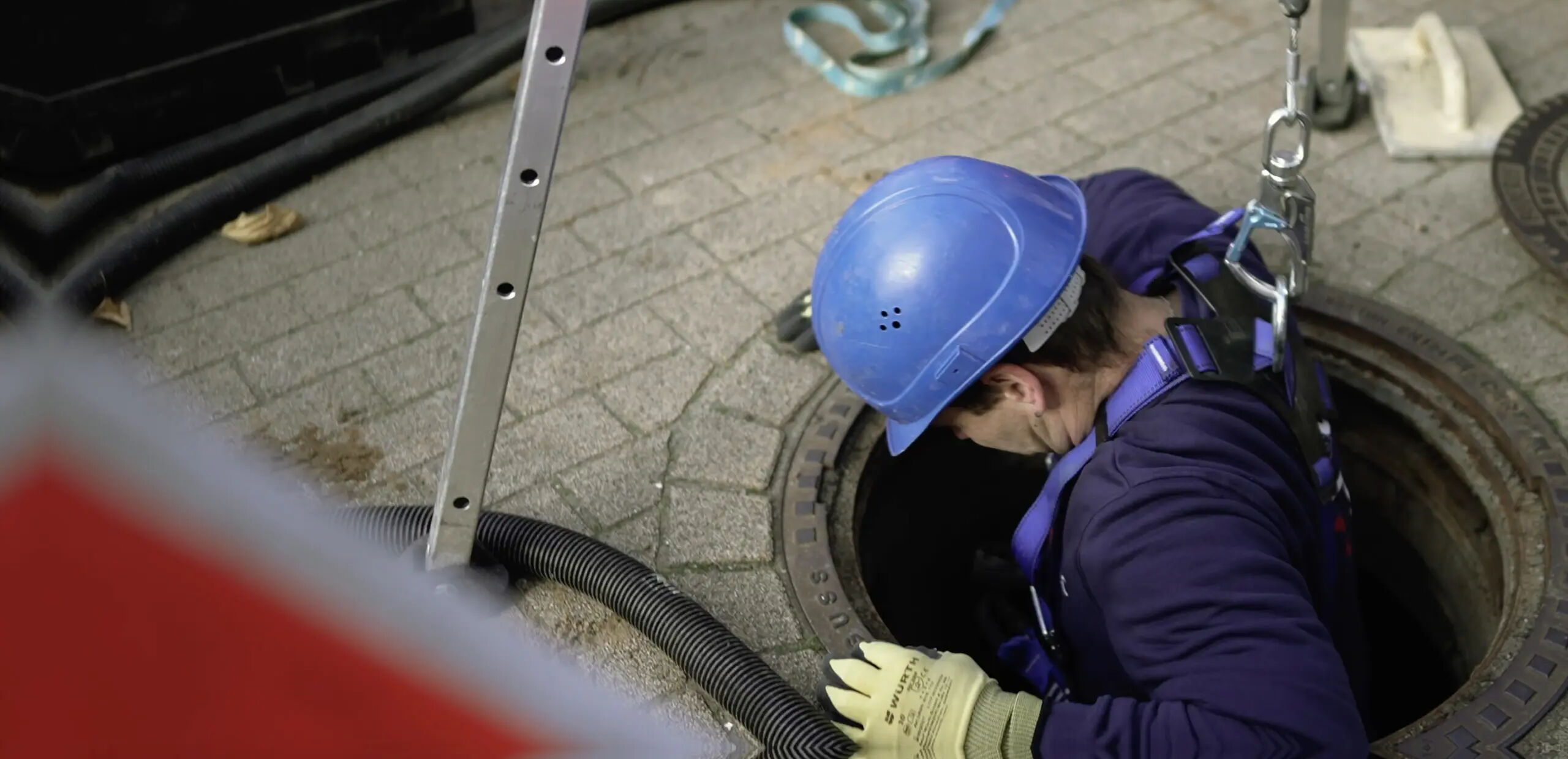 Eine Person in Sicherheitskleidung mit blauem Helm und Handschuhen steigt in einen Gullyschacht. Eine Leiter steht daneben, und einige Ausrüstungsgegenstände liegen auf dem gepflasterten Boden in der Nähe.