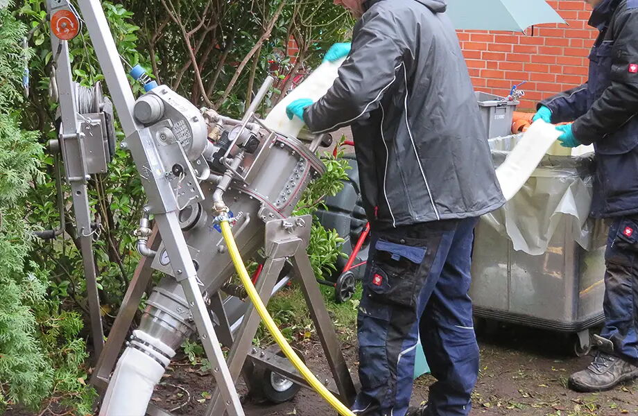 Zwei Personen in Arbeitskleidung bedienen eine Maschine im Freien. Die Maschine hat ein Metallgestell, an das ein Schlauch und Kabel angeschlossen sind. Die Umgebung ist grün mit Pflanzen und einem roten Backsteingebäude im Hintergrund.
