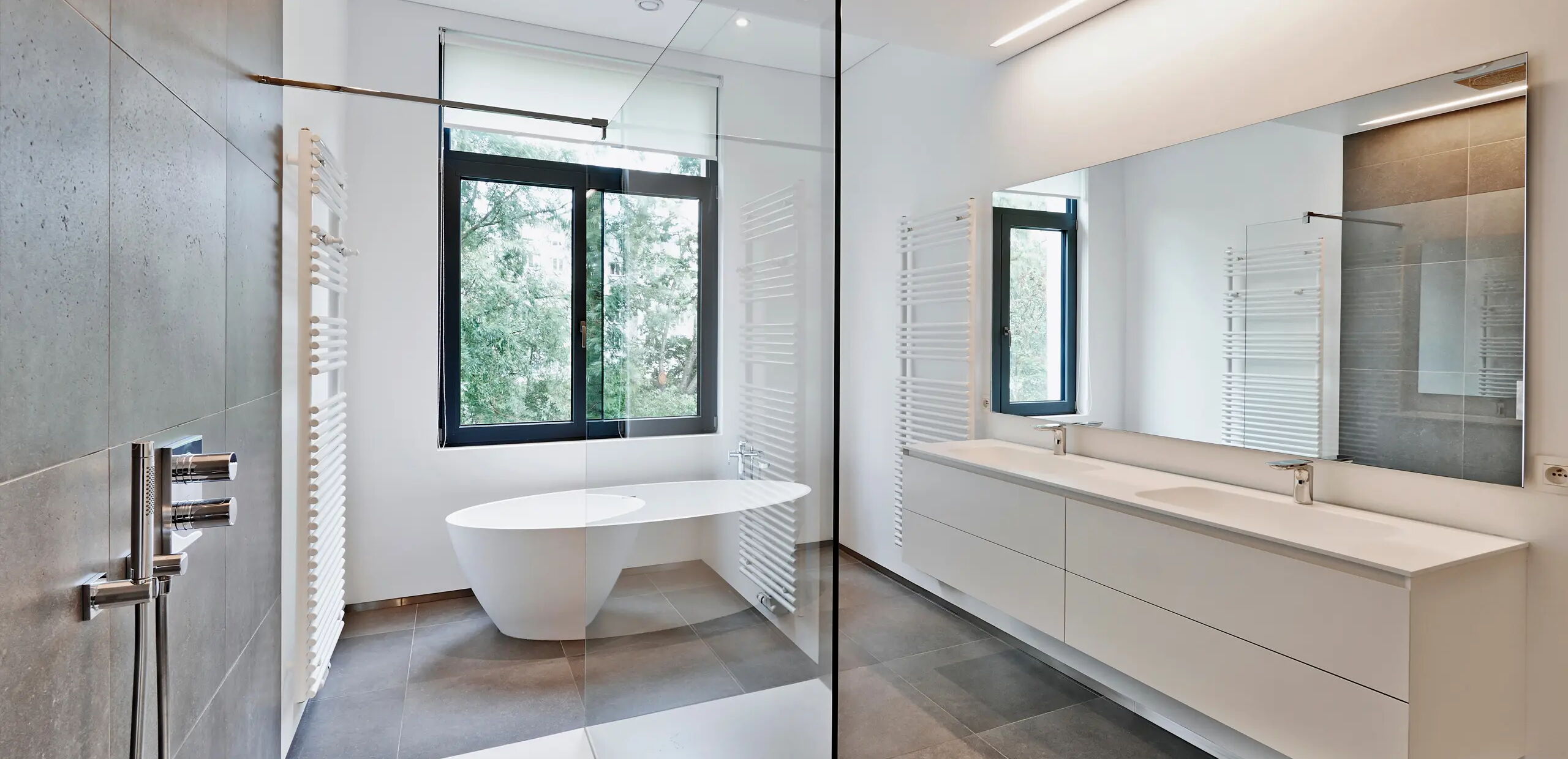 Modernes Badezimmer mit großen Fenstern, die Tageslicht hereinlassen. Eine freistehende Badewanne steht vor den Fenstern. Wandmontierte Waschbecken auf einem langen, weißen Waschtisch. Gläserne Duschabtrennung und Handtuchheizkörper.