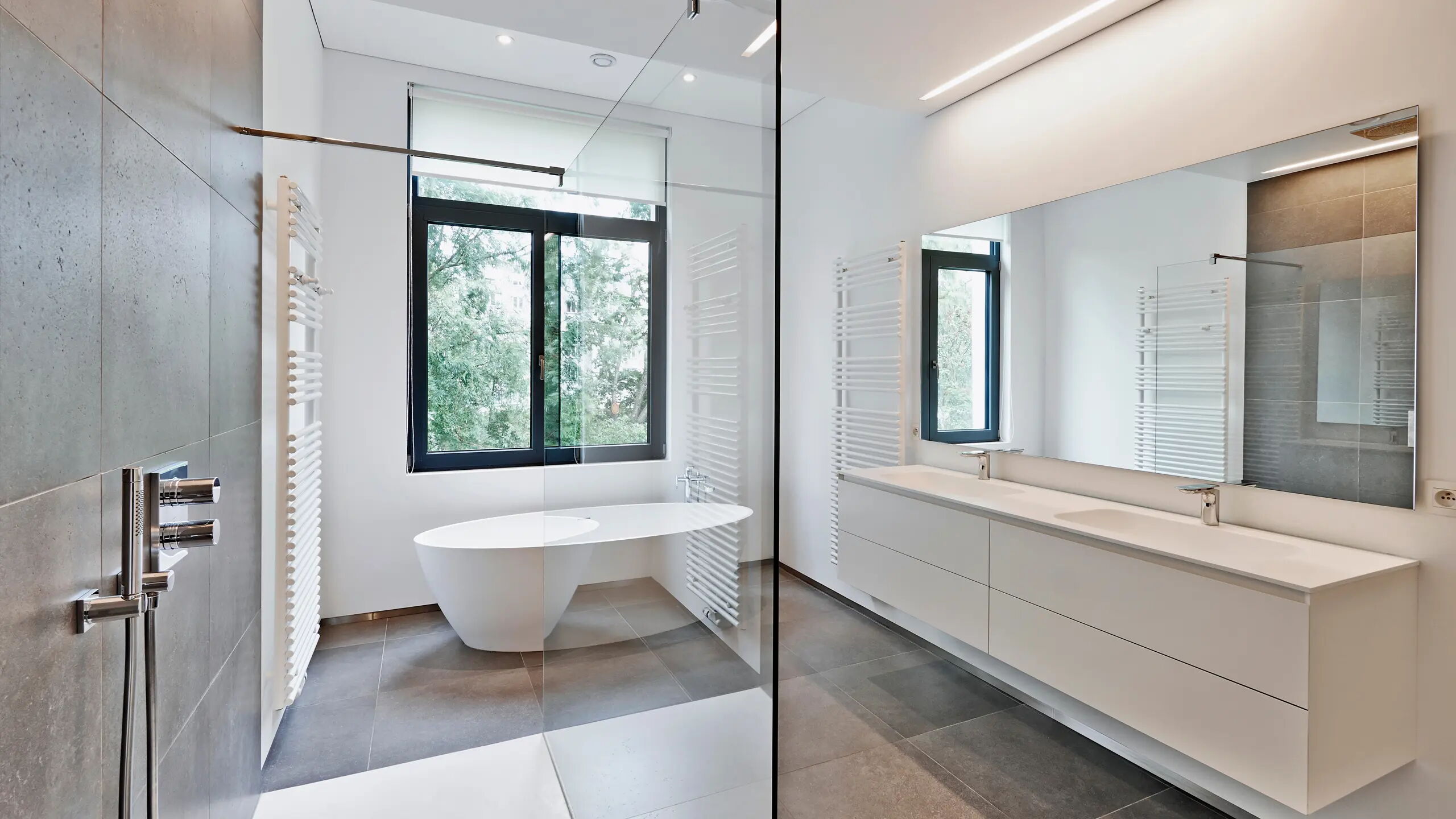 Modernes Badezimmer mit großen Fenstern, die Tageslicht hereinlassen. Eine freistehende Badewanne steht vor den Fenstern. Wandmontierte Waschbecken auf einem langen, weißen Waschtisch. Gläserne Duschabtrennung und Handtuchheizkörper.