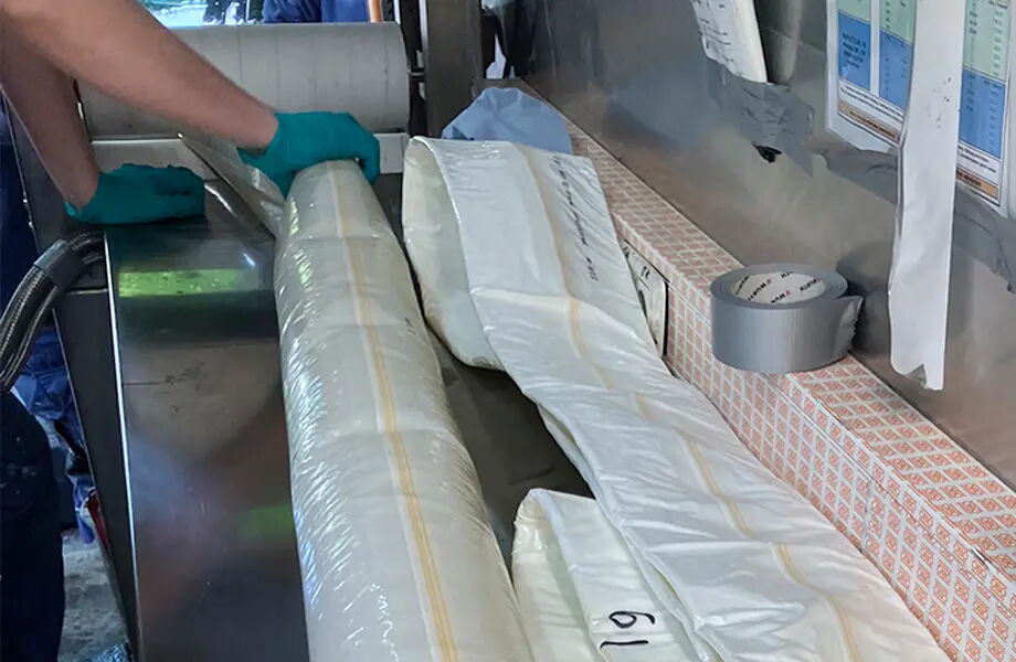 Ein Mensch mit blauen Handschuhen hält eine Rolle weißer Plastikfolie. Daneben liegen weitere Folienrollen auf einem Tisch, auf dem auch Klebeband und Listen zu sehen sind.