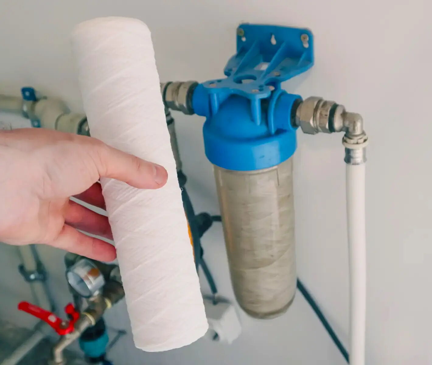 Eine Hand hält einen weißen, zylinderförmigen Wasserfilter in der Nähe eines installierten blauen Filtersystems an einer Wand. Die Rohre sind mit dem Filtersystem verbunden.