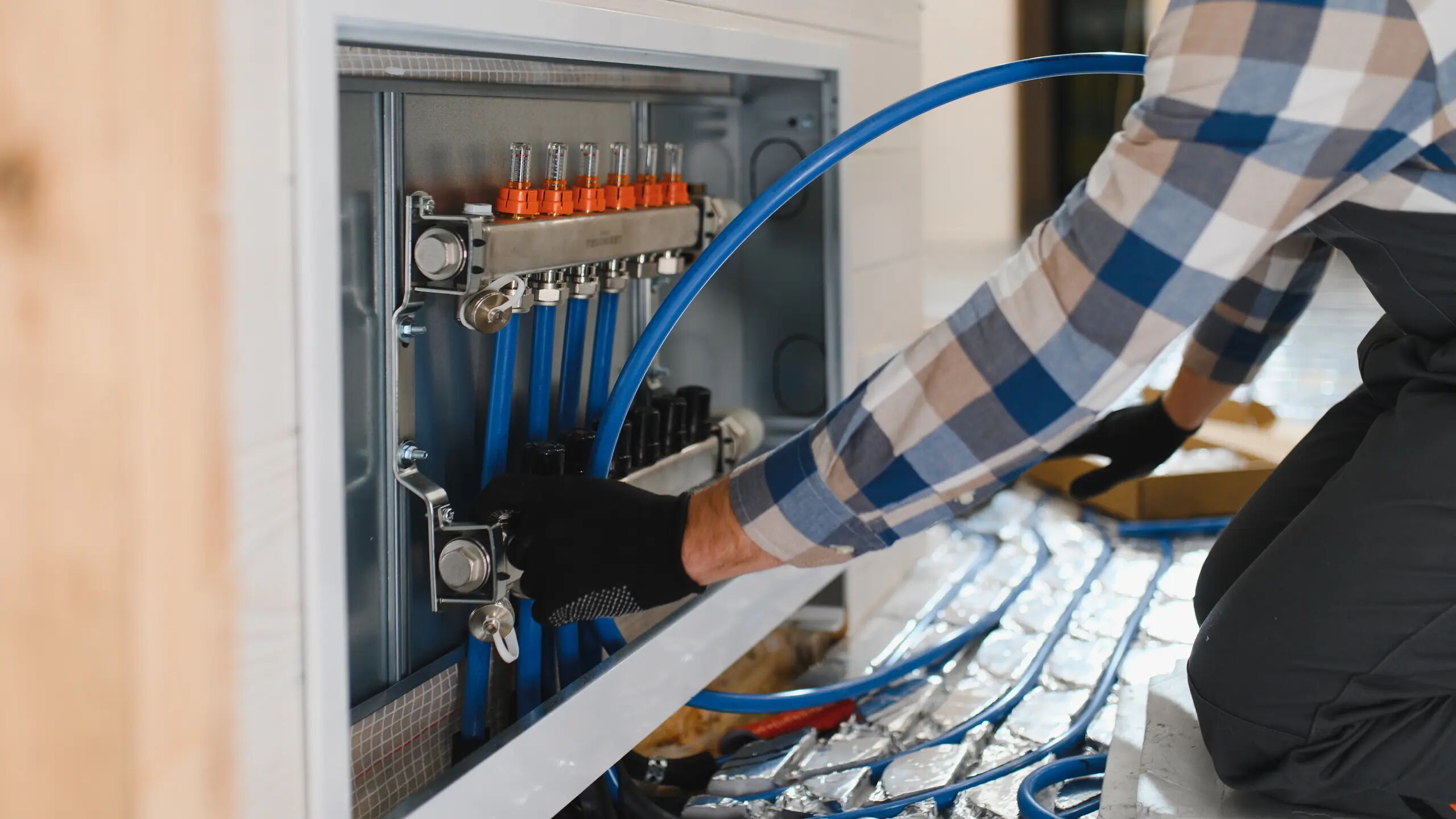 Eine Person mit kariertem Hemd und schwarzen Handschuhen arbeitet an einer Heizungsinstallation. Blaue Röhren sind an eine Verteilungseinheit angeschlossen. Der Boden ist mit reflektierenden Materialien bedeckt.
